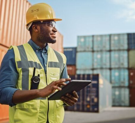 Smiling Portrait of a Handsome African American Black Industrial Engineer in Yellow Hard Hat and Safety Vest Working on Tablet Computer. Foreman or Supervisor in Container Terminal.